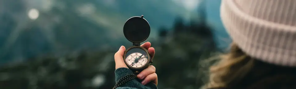 A woman in a pink toboggan hat holds up a bronze compass against a distant background of trees and mountains.