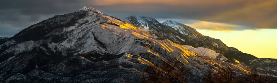 The Wasatch Mountains, covered in snow and brown trees, highlighted on the right by a twilight sun that lights up the slopes in yellow and gold.